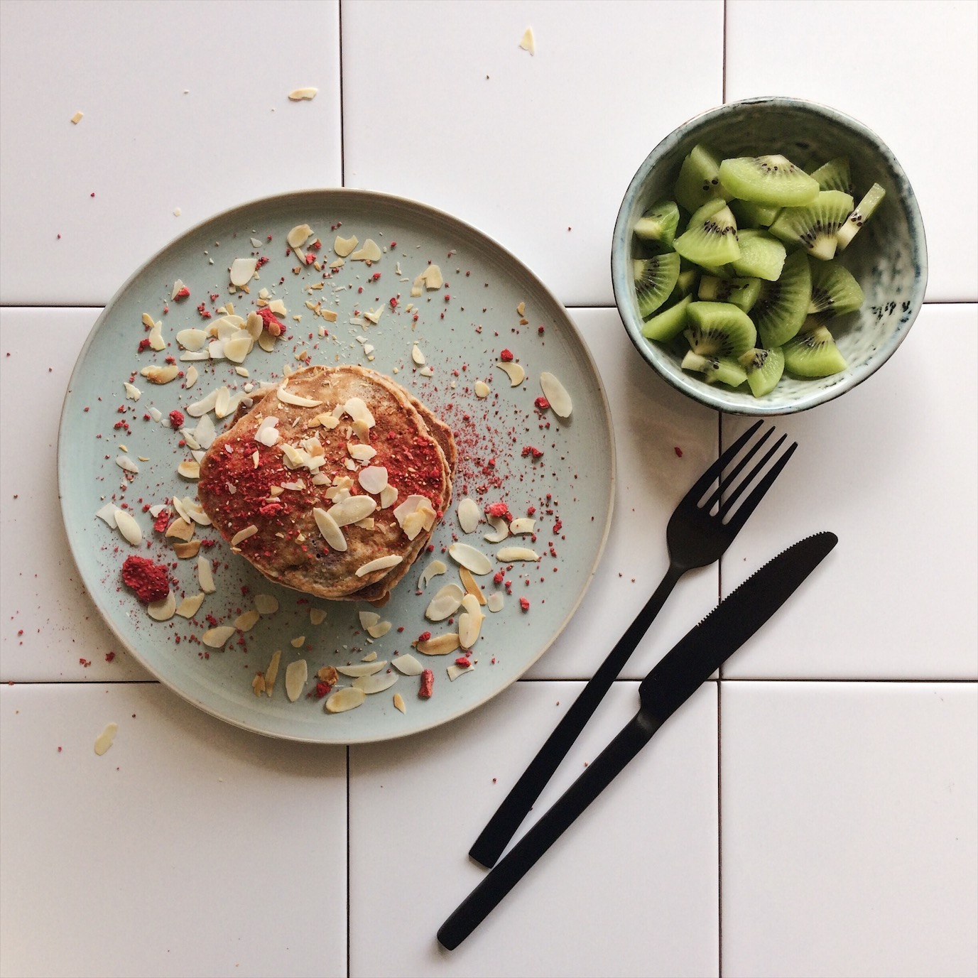 Flatlay van American pancakes versierd met amandelschaafsel en gedroogde aarbeien met daarnaast een bakje met stukjes kiwi