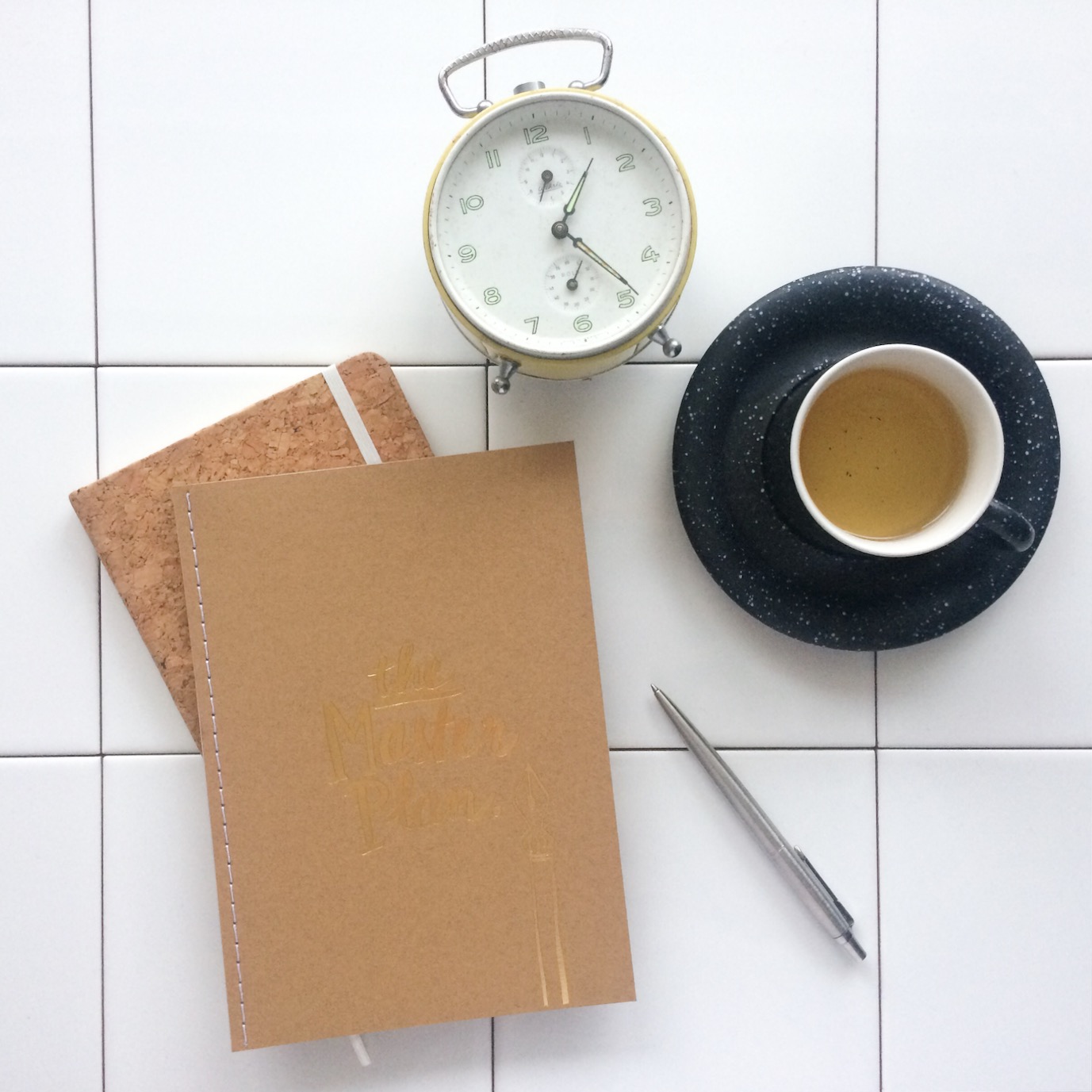 Flatlay van notitieboek met 'the masterplan' erop, lichtgele wekker op pootjes, pen en theekop met schotel