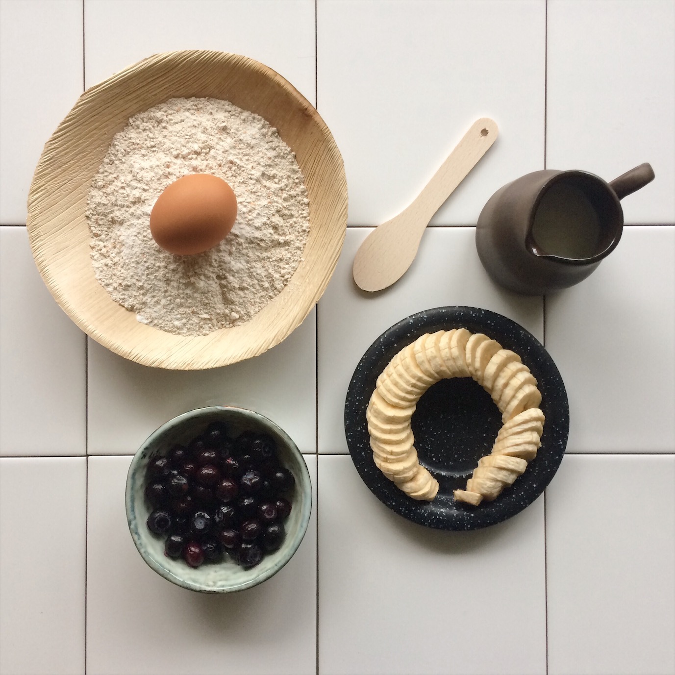 Flatlay van de ingredienten voor bananen pancakes met blauwe bessen: volkoren speltmeel, eieren, banaan, blauwe bessen en melk